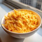 Close-up of a white bowl filled with creamy, melted cheese topped with extra shredded cheddar for beer cheese dip.