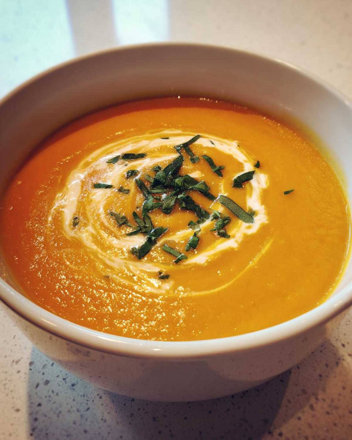 Close-up of a bowl of vibrant orange butternut squash soup topped with cream swirl and fresh herbs.