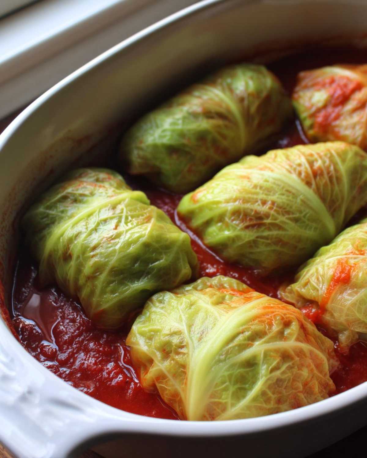 Close-up of several stuffed cabbage rolls simmering in a rich tomato sauce inside a white baking dish.