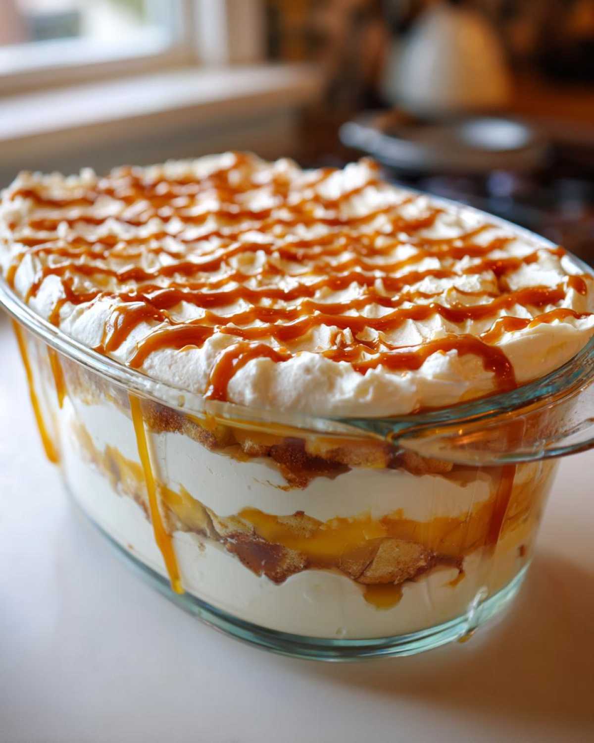 A glass dish filled with layers of cream, cake/cookies, and caramel sauce, topped with whipped cream and drizzled caramel for a caramel apple trifle.
