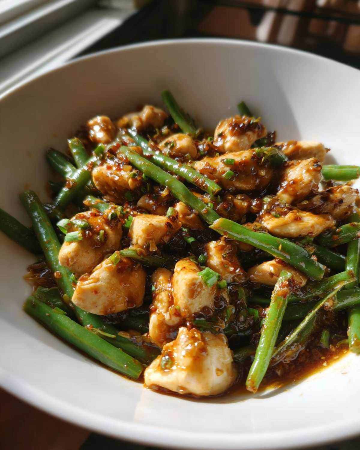 Close-up of a white bowl filled with saucy chicken and green beans, garnished with chives.