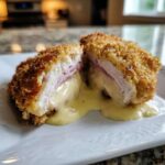 Cross-section of crispy chicken cordon bleu showing melted cheese and ham oozing out onto a white plate.