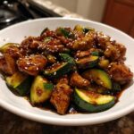 A white bowl filled with glossy chicken zucchini stir fry, topped with white sesame seeds.