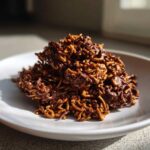A mound of rich, chocolate-coated haystack cookies made with chow mein noodles on a white plate.