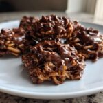 A close-up shot of several glossy, chocolate-covered haystack cookies piled on a white plate.