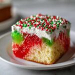 A moist slice of christmas poke cake featuring layers of red and green jello, whipped topping, and festive sprinkles.