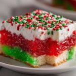 Close-up of a slice of Christmas poke cake featuring layers of green and red jello, white whipped topping, and holiday sprinkles.