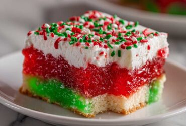 Close-up of a slice of Christmas poke cake featuring layers of green and red jello, white whipped topping, and holiday sprinkles.