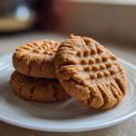 Three classic peanut butter cookies stacked on a white plate, showing the traditional criss-cross fork marks.