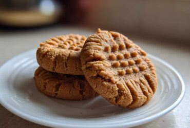 Three classic peanut butter cookies stacked on a white plate, showing the traditional criss-cross fork marks.