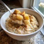 A spoonful of creamy apple cinnamon oatmeal topped with diced apples and cinnamon being lifted from a white bowl.
