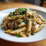 A close-up of a white plate piled high with creamy beef pasta, featuring penne coated in a rich sauce and topped with fresh parsley.