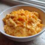 A small white bowl filled with thick, creamy, orange beer cheese dip, topped with shredded cheddar cheese.
