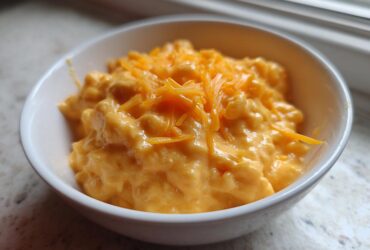 A small white bowl filled with thick, creamy, orange beer cheese dip, topped with shredded cheddar cheese.