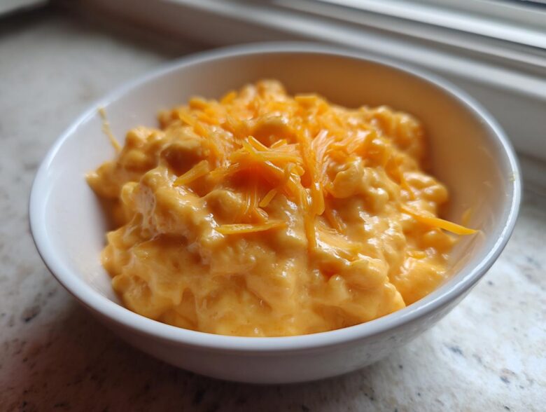 A small white bowl filled with thick, creamy, orange beer cheese dip, topped with shredded cheddar cheese.