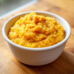 A close-up of a white bowl filled with rich, creamy, yellow cheese grits sitting on a wooden surface.