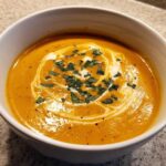A close-up of creamy, vibrant orange butternut squash soup topped with a swirl of cream and fresh herbs.