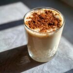 A glass of creamy, light-colored coquito topped generously with ground cinnamon.