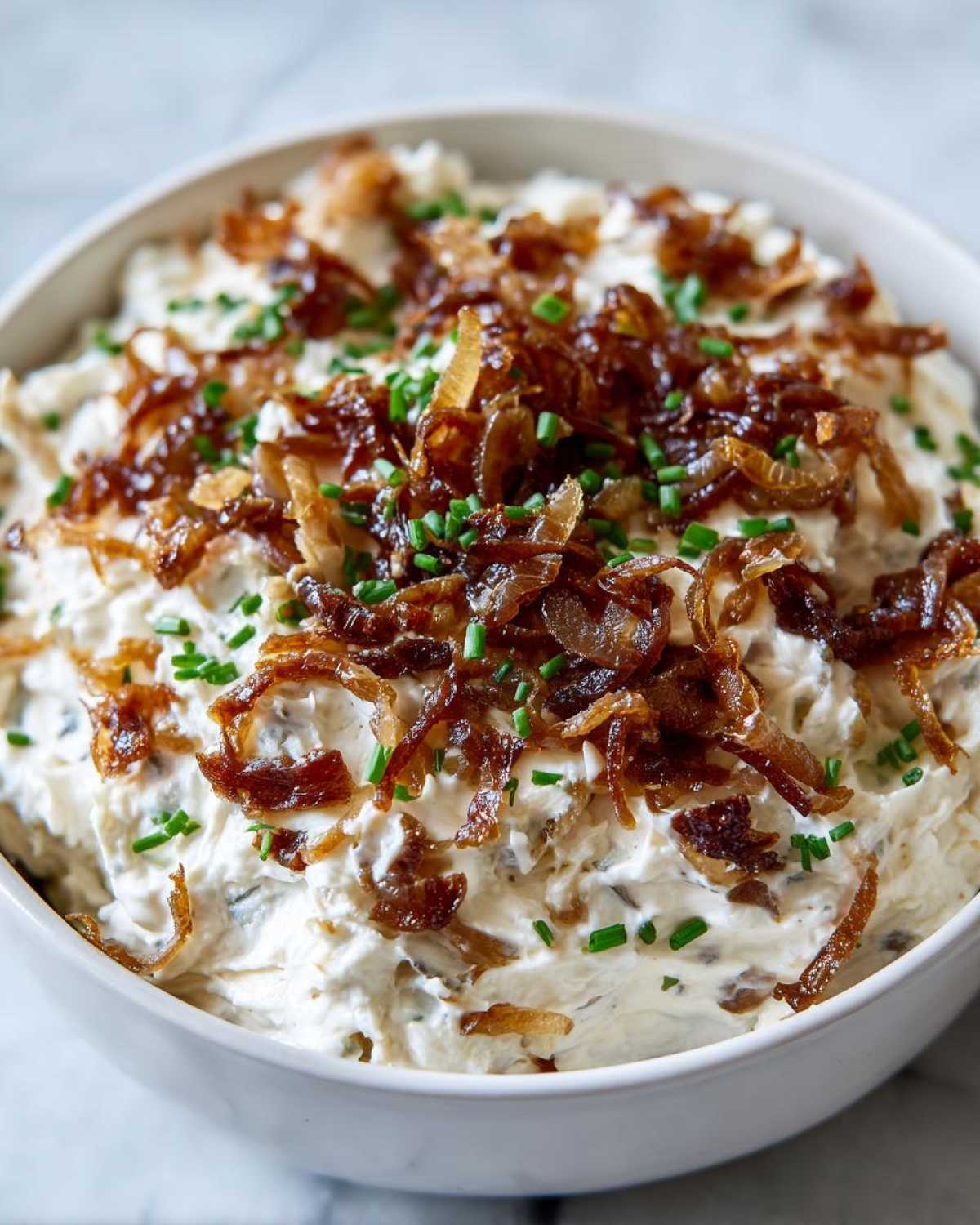 A close-up of a creamy homemade onion dip topped generously with crispy fried onions and fresh chives.