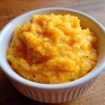 Close-up of rich, creamy, orange-colored cheese grits served in a small white fluted ramekin on a wooden surface.