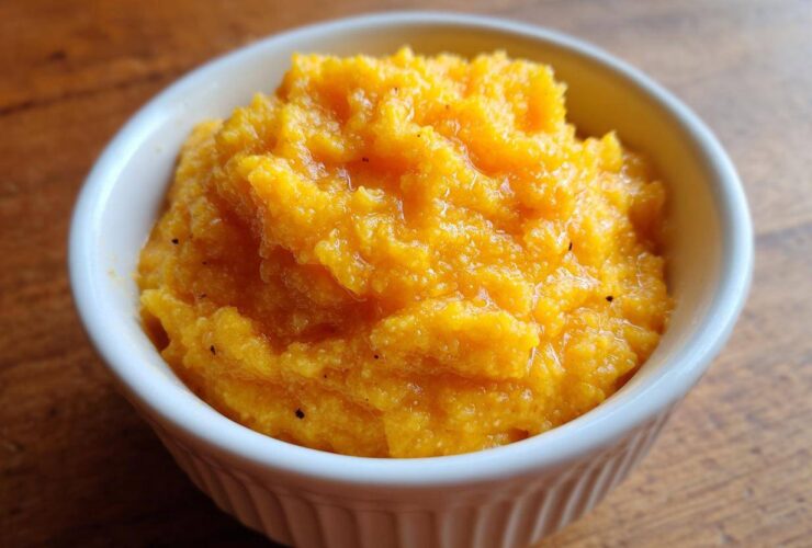 Close-up of rich, creamy, orange-colored cheese grits served in a small white fluted ramekin on a wooden surface.