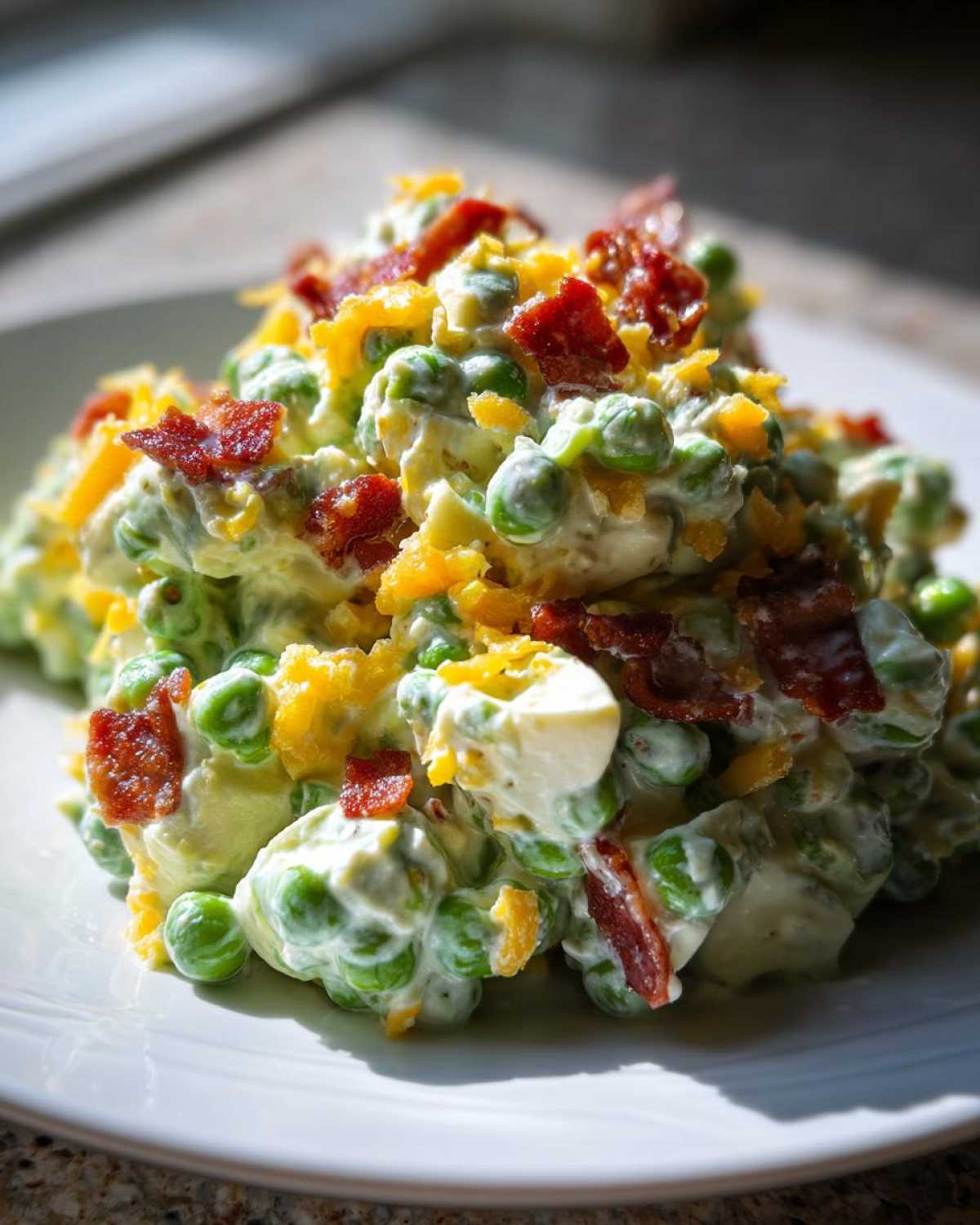 A close-up of creamy pea salad loaded with green peas, covered in a white dressing, topped with crumbled bacon and shredded cheddar cheese.