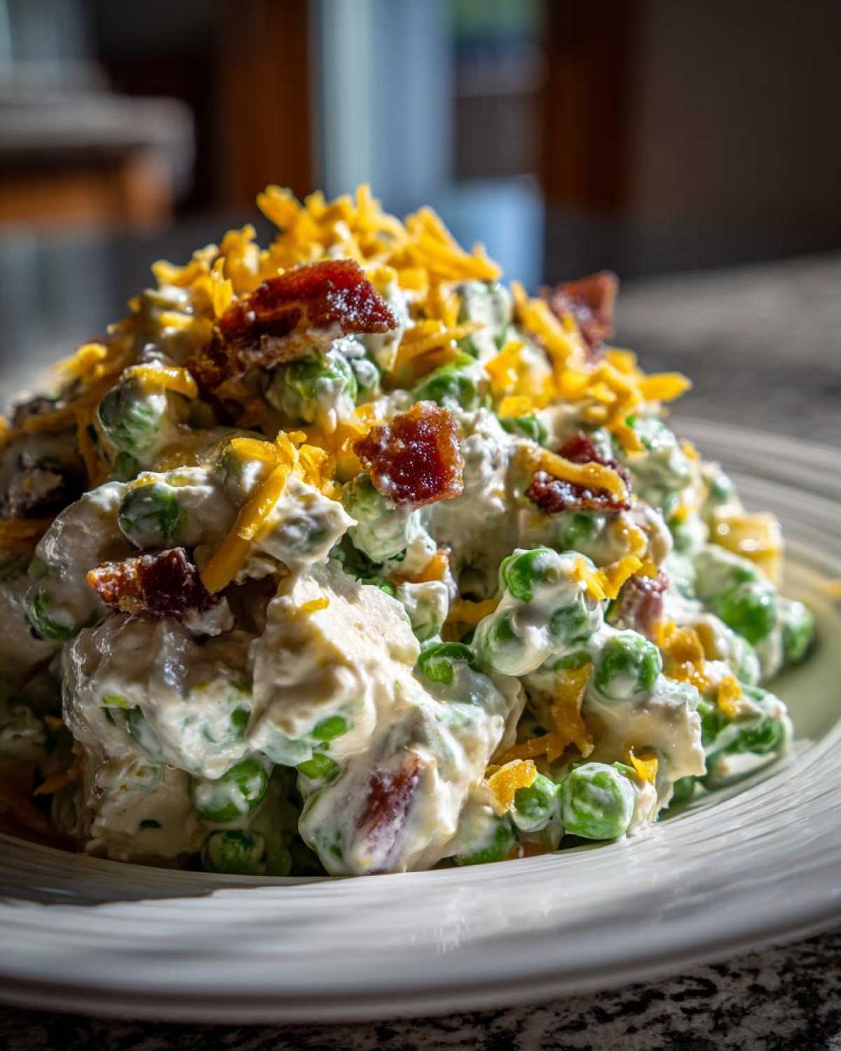 Close-up of creamy pea salad mixed with a white dressing, topped generously with shredded cheddar cheese and crispy bacon pieces.