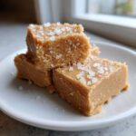 Three squares of creamy peanut butter fudge topped with coarse sea salt flakes on a white plate.