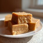Close-up of creamy peanut butter fudge squares stacked on a small white plate, catching bright sunlight.