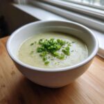 A close-up of creamy potato leek soup in a white bowl, garnished generously with chopped green chives and pepper.