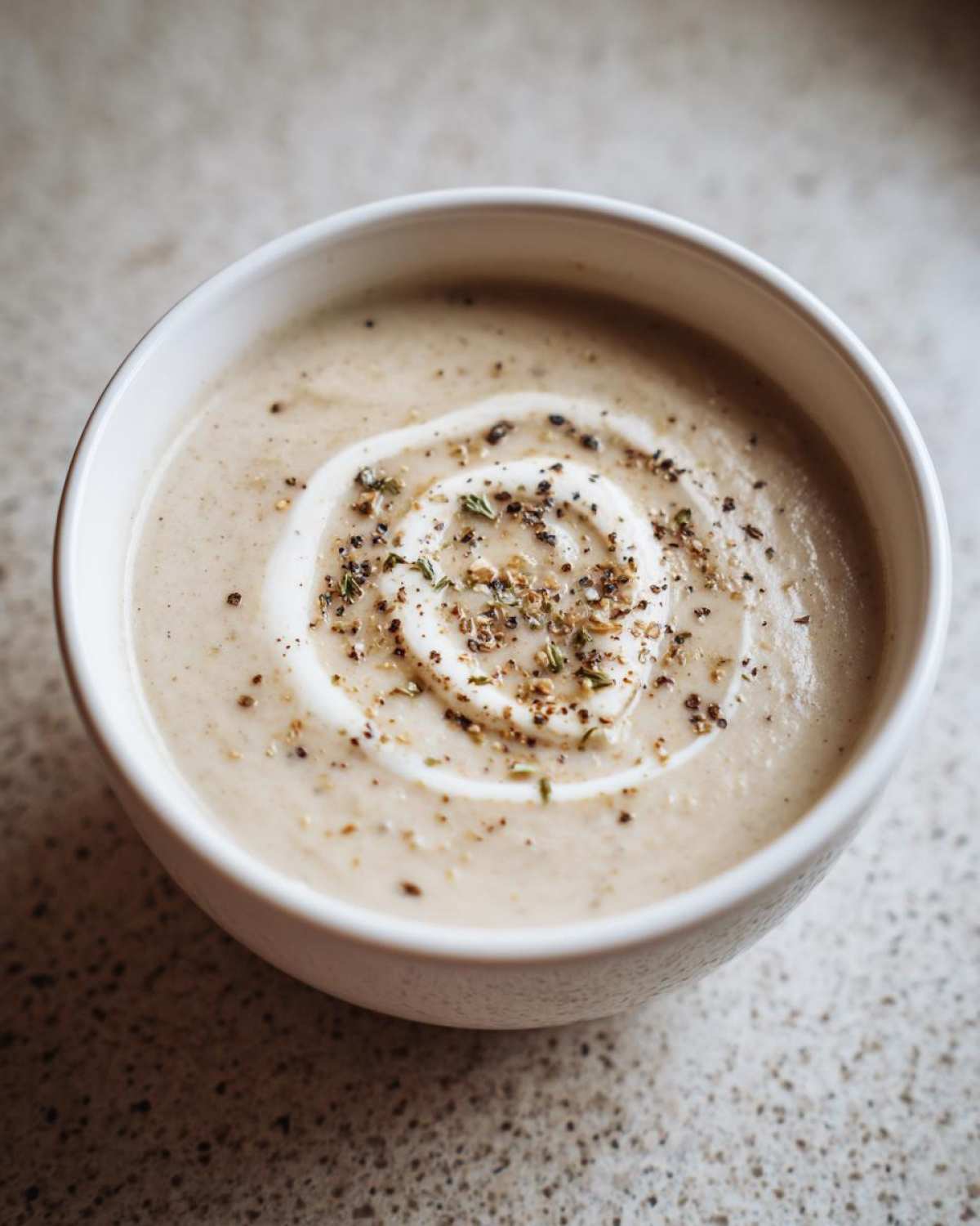 A close-up of a bowl of creamy roasted cauliflower soup, garnished with a swirl of cream and cracked pepper.