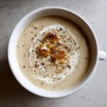 Overhead view of creamy roasted cauliflower soup topped with a swirl of cream, cracked pepper, and roasted cauliflower florets.