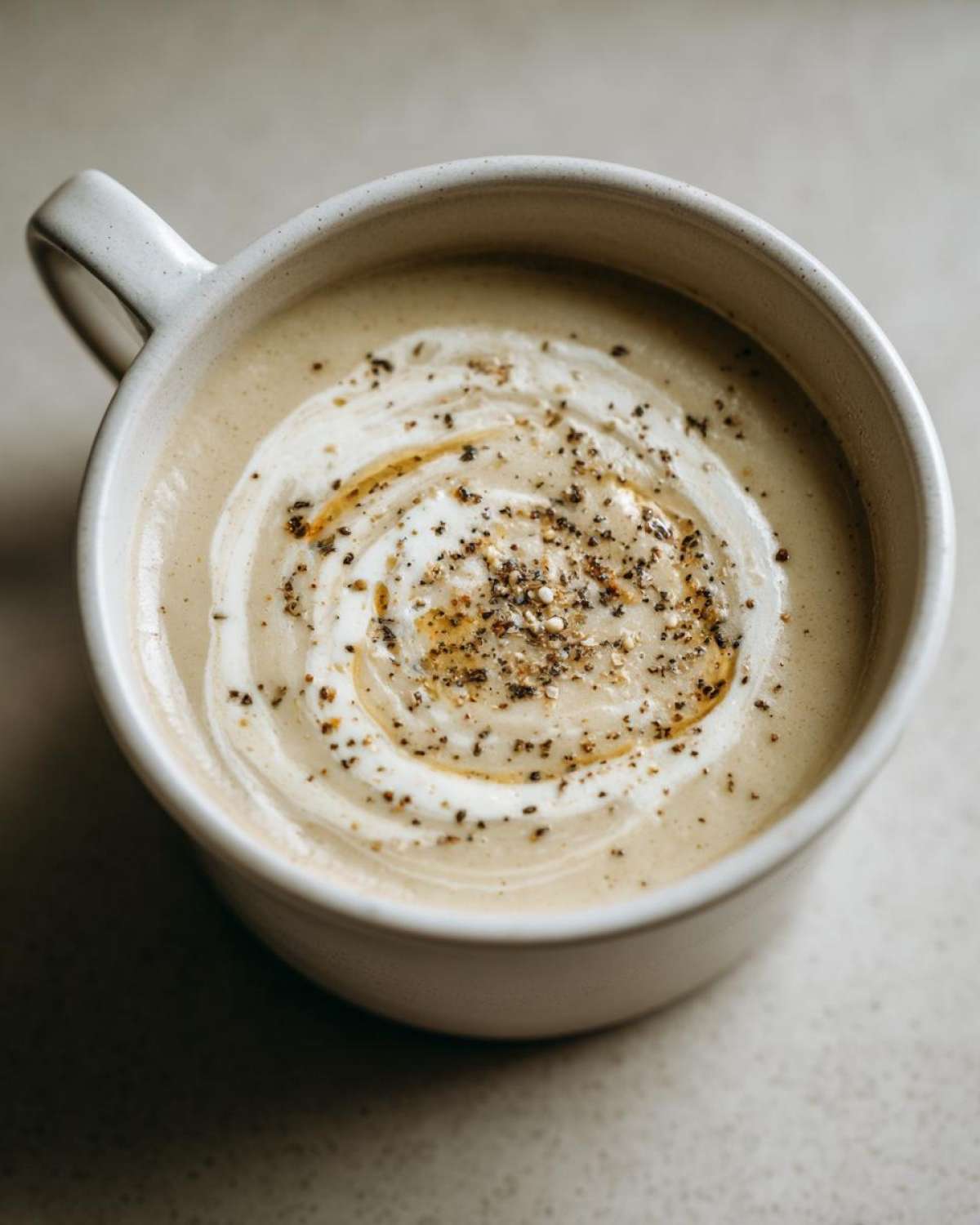 Overhead close-up of creamy roasted cauliflower soup garnished with a swirl of cream and cracked black pepper.