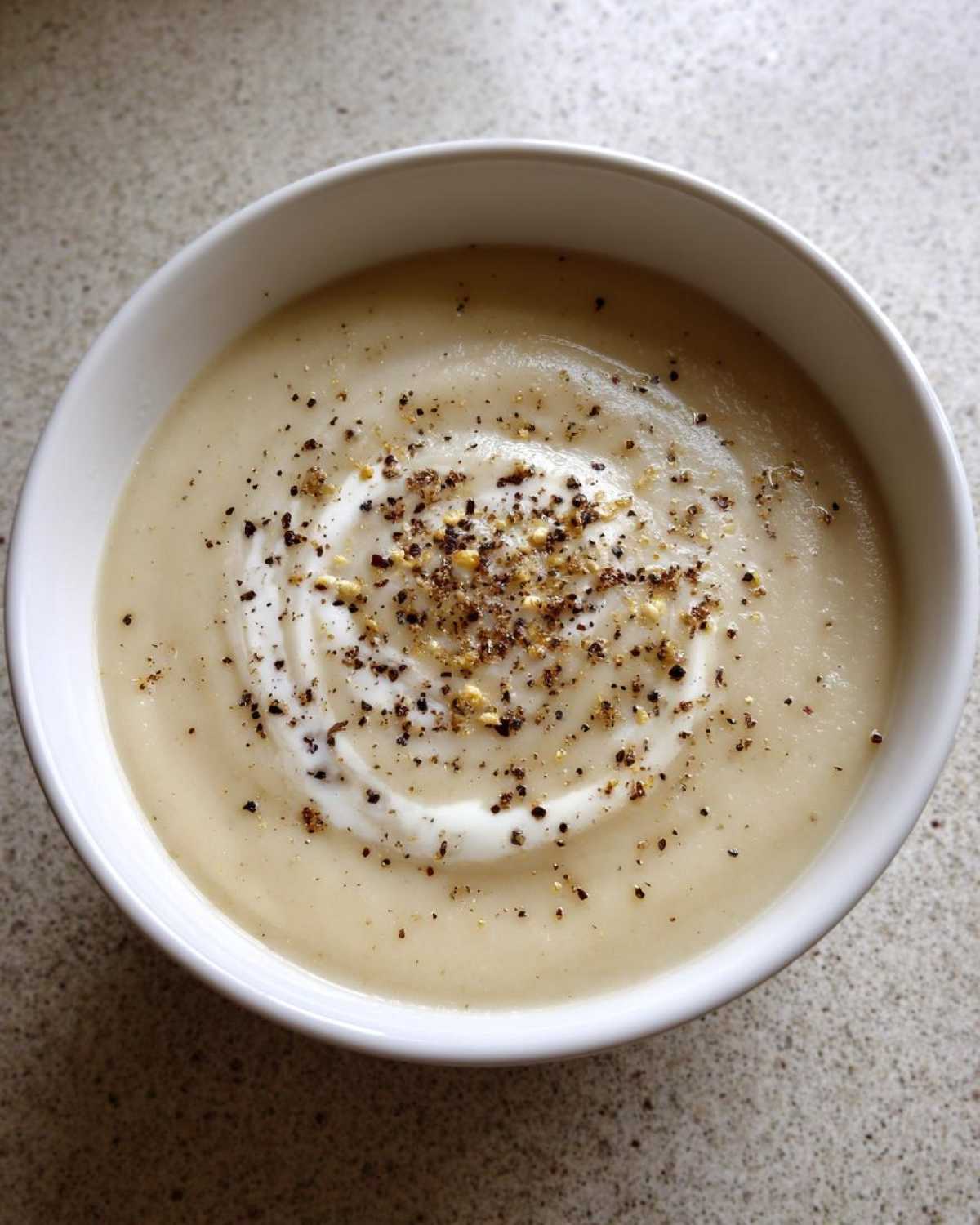 A close-up of creamy roasted cauliflower soup, swirled with cream and topped generously with cracked black pepper.
