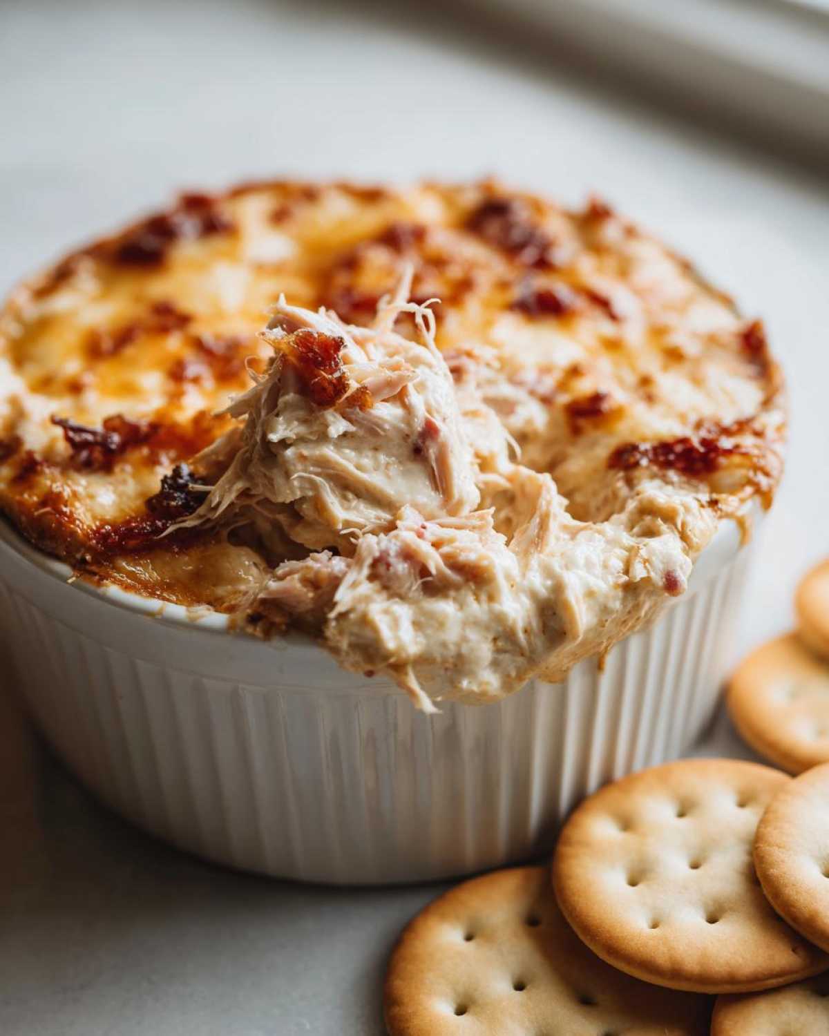 A spoonful of creamy, shredded reuben dip being lifted from a white ramekin next to round crackers.