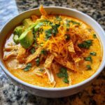 Close-up of a bowl of creamy, orange-hued tortilla soup topped with shredded cheese, avocado, and cilantro.