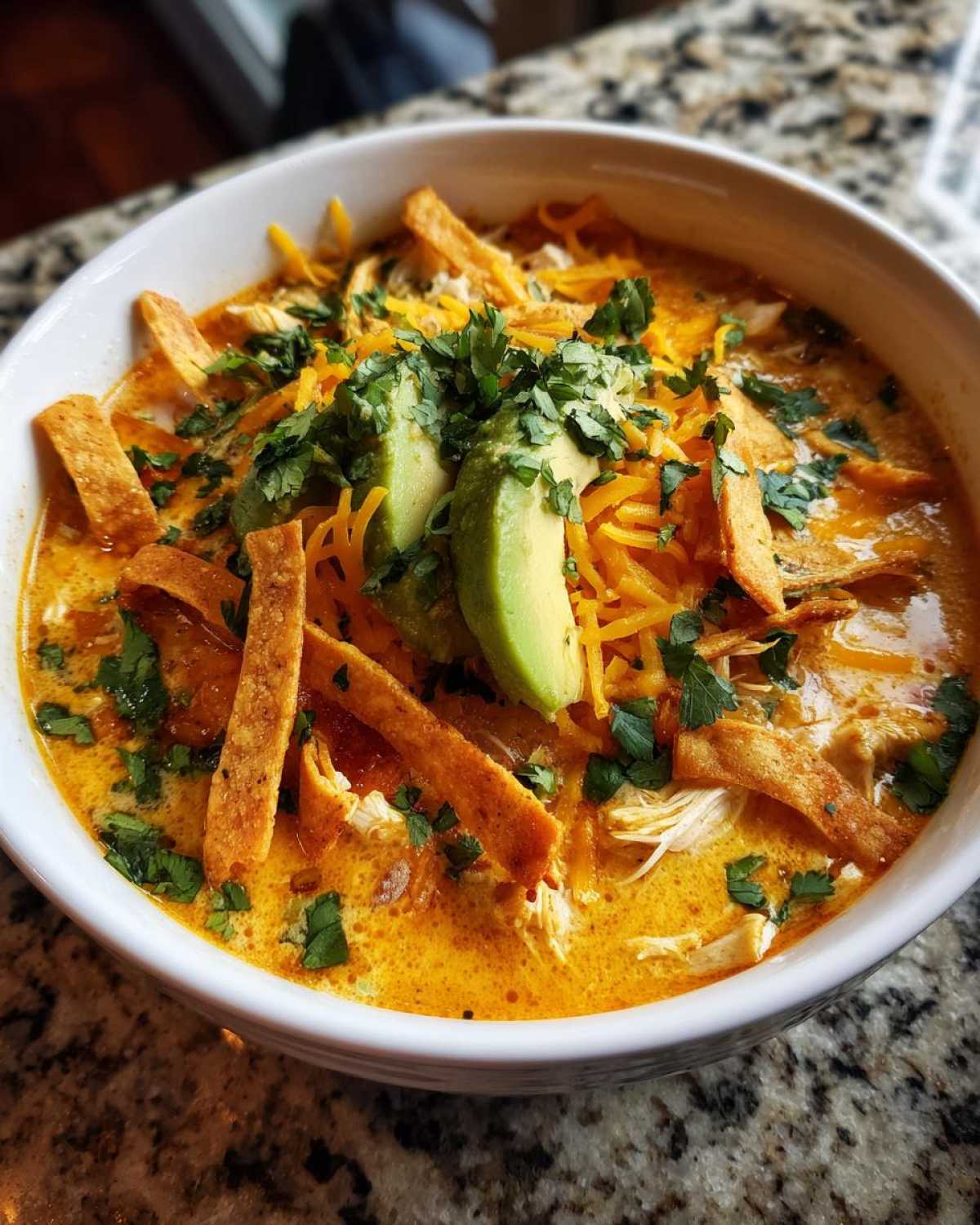 A close-up of creamy tortilla soup topped with shredded cheese, cilantro, avocado slices, and tortilla strips.