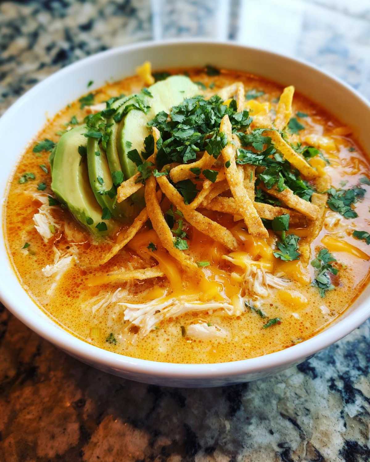 A close-up of a bowl of creamy tortilla soup, generously topped with sliced avocado, shredded cheese, crispy tortilla strips, and cilantro.