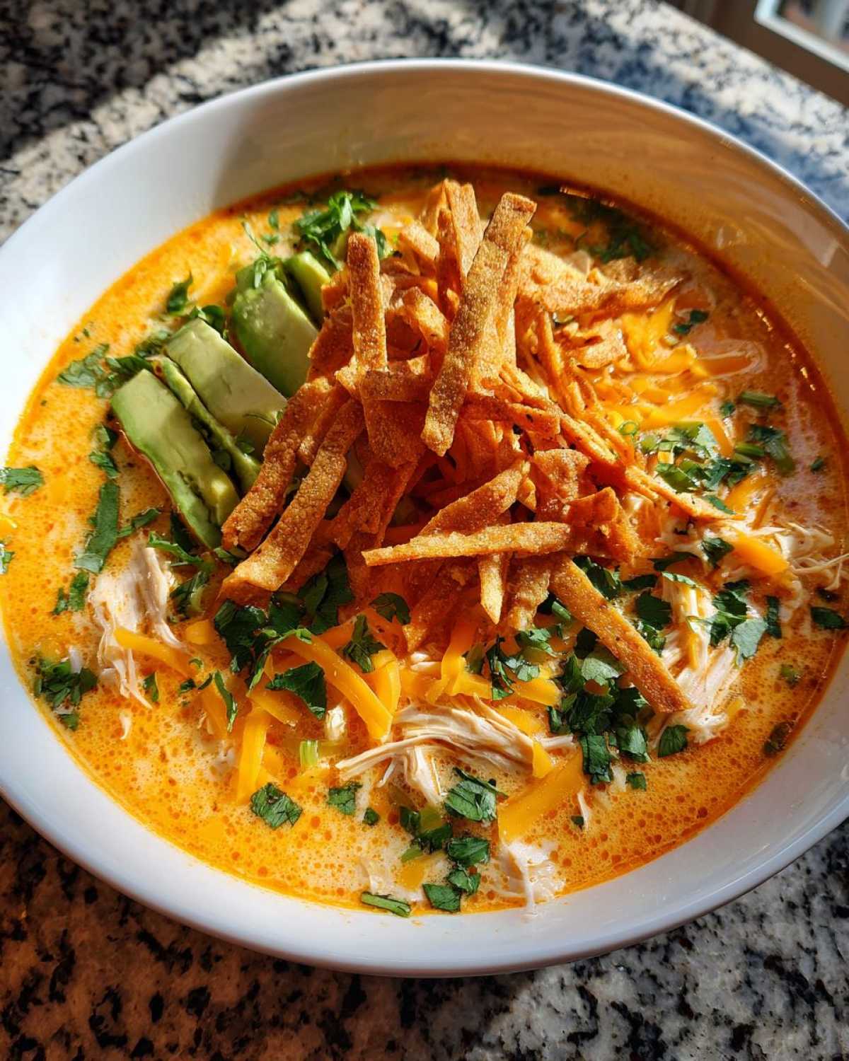 A close-up of creamy tortilla soup topped with shredded chicken, avocado slices, cheese, and crispy tortilla strips.