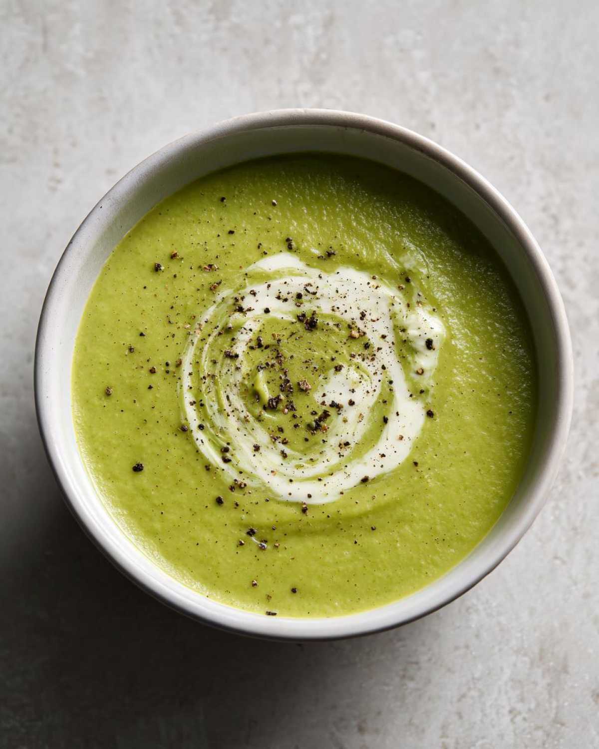 Overhead view of a bowl of vibrant green creamy zucchini soup, swirled with cream and topped with cracked black pepper.