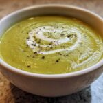 Close-up of a bowl of vibrant green, creamy zucchini soup topped with a swirl of cream and cracked black pepper.