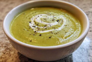 Close-up of a bowl of vibrant green, creamy zucchini soup topped with a swirl of cream and cracked black pepper.