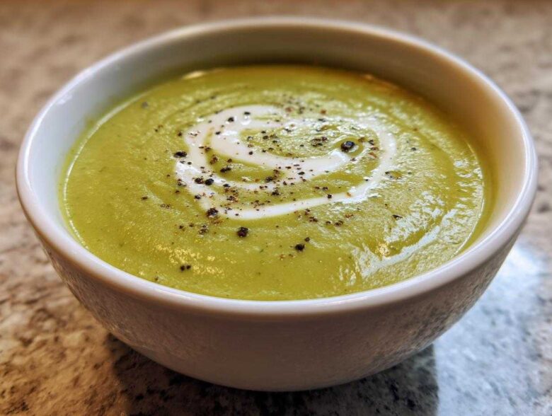 Close-up of a bowl of vibrant green, creamy zucchini soup topped with a swirl of cream and cracked black pepper.