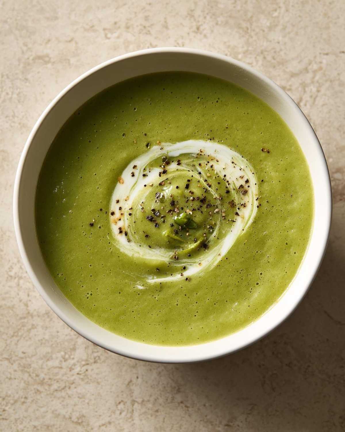 Overhead view of a bowl of vibrant green, creamy zucchini soup topped with a swirl of cream and cracked black pepper.