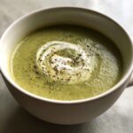 A close-up of creamy zucchini soup in a white bowl, topped with a swirl of cream and cracked black pepper.