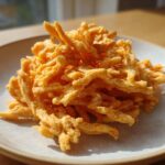 A mound of golden, crispy cheese straws piled high on a light-colored ceramic plate, catching bright sunlight.