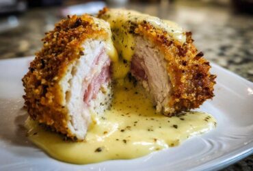 A cross-section of crispy, breaded chicken cordon bleu revealing ham and cheese, drizzled with a creamy herb sauce.