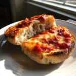 Two halves of a freshly made English muffin pizza, topped with melted cheese and sauce, sitting on a white plate.