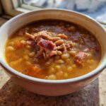 Close-up of a steaming white bowl filled with hearty bean soup topped with shredded ham.