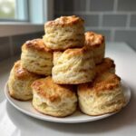 A tall stack of golden-brown, flaky buttermilk biscuits piled high on a white plate.